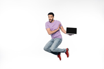 Fototapeta na wymiar amazed man in jeans and purple t-shirt levitating while holding laptop on white.