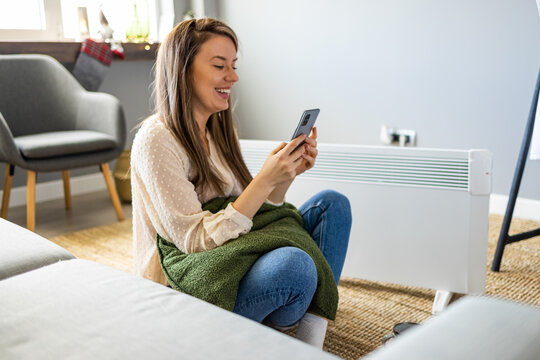 Young woman in sweater feeling cold, waiting for the house to heat up, controlling heating system with a smart home thermostat. Woman and radiator using smartphone app for temperature control