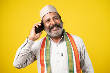 smiling Seniot Indian politician busy talking on mobile phone on yellow studio background - concept of happiness, positive emotion and communication skills - obrazy, fototapety, plakaty