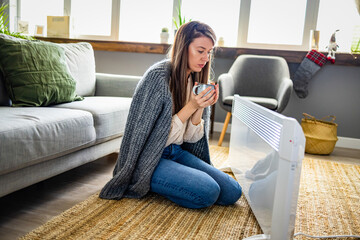 Obraz premium Woman in living room, focus on electric fan heater. Using heater at home in winter. Woman warming her hands. Heating season. Cold adult woman covered with clothes at home