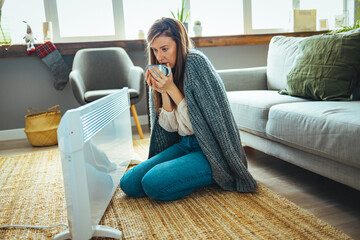 Woman in living room, focus on electric fan heater. Using heater at home in winter. Woman warming her hands. Heating season. Cold adult woman covered with clothes at home