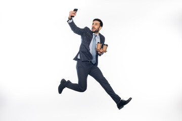 cheerful businessman in suit jumping while holding cup holder with paper cups and taking selfie on white.