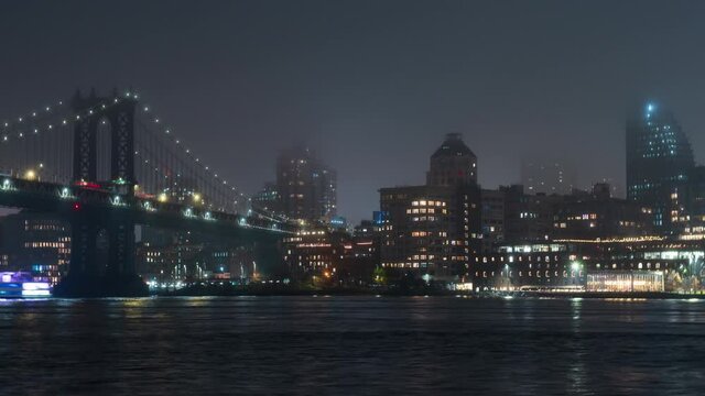 Zooming out timelapse of the high buildings of Brooklyn district, illuminated Brooklyn bridge that crosses the East River in New York City, USA. Night. Touristic ferries sail along the river