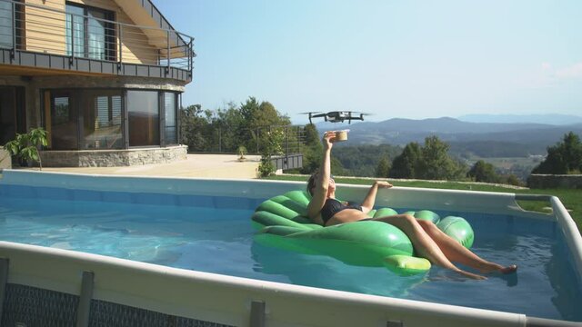 COPY SPACE: Young woman lounging in a pool gets her food delivered by a contactless UAV service. Food delivery drone brings a brown disposable bowl to a Caucasian woman relaxing in her home pool.