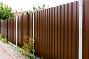 Close-up of a brown metal profile fence. Corrugated surface. Copy space. Security. Private property fencing. Opaque hedge. Outdoor house exterior. Steel material. High. Side view. Urban or indastrial.
