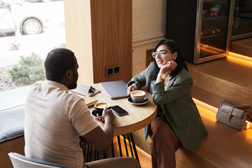 Portrait of two young people chatting at table during coffee break in graphic cafe interior, copy...