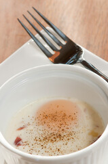 top view, close distance of a pouched  egg in a white ramekin, on square, white plate and artisan fork, sprinkled with black pepper  