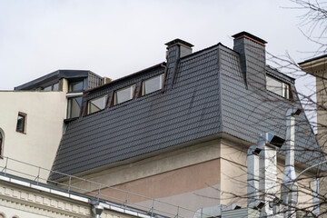 Fototapeta na wymiar Sloping roof with metal coating of residential building with windows. Roof with drains, slopes, rain pipes and gutters, tides and dormer windows against sky