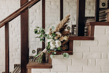 Fototapeta na wymiar A beautiful boho style bridal bouquet is attached to a wooden railing, which is part of the staircase, inside the house. A wedding bouquet in a boho style lies against the background of a white brick 