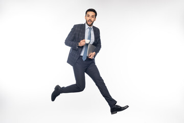 excited businessman in suit jumping while holding laptop and cup on white.