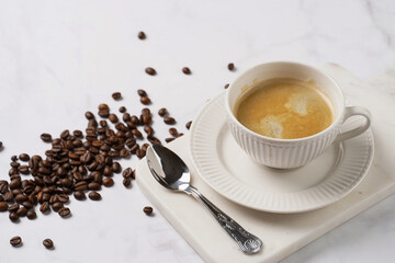 a white porcelain cup with coffee with milk on a marble board on a white table surrounded by scattered coffee beans