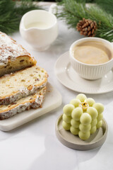 White porcelain cup with coffee cappuccino on a white saucer plate, christmas pastry stollen and a pastel green bubble candle on a marble table