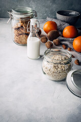 frascos de cristal con avena, leche y copos de avena, con un puñado de frutos secos, naranjas y kiwis, sobre una mesa gris claro