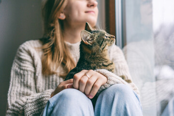 Young cheerful girl sitting at home on the windowsill in a warm sweater playing with a gray cat on a winter day