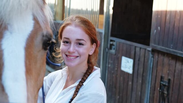Young female rider standing by horse in stable, smiling