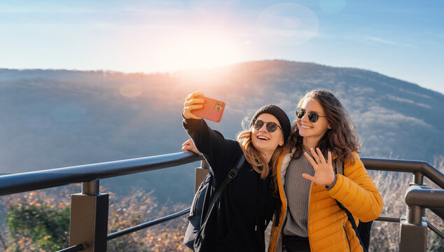 Two happy cheerful young girls in sunglasses traveling together taking selfie on smartphone with mountain view - Powered by Adobe