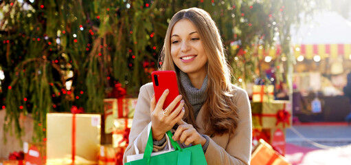 Fashion stylish woman receiving christmas wishes on smartphone when walking between christmas markets outdoors