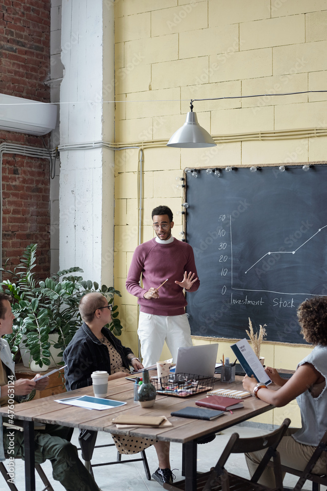 Poster young confident businessman explaining financial graph to colleagues at seminar while standing by bl
