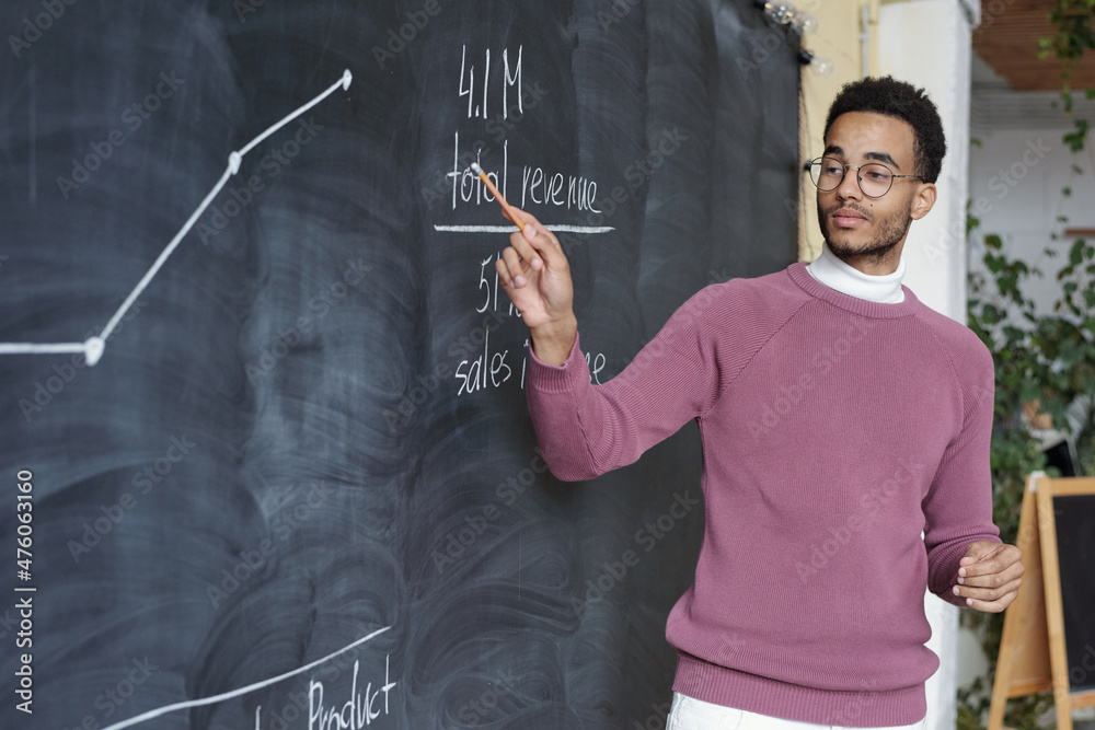 Poster young confident teacher of economics pointing at graph on blackboard while explaining it to colleagu