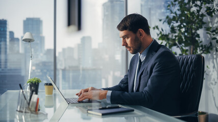 Confident Businessman in a Suit Sitting at a Desk in Modern Office, Using Laptop Computer, Next to Window with Big City with Skyscrapers View. Successful Finance Manager Planning Work Projects.