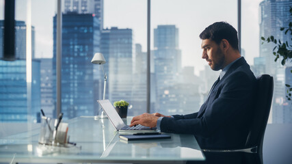 Confident Businessman in a Suit Sitting at a Desk in Modern Office, Using Laptop Computer, Next to Window with Big City with Skyscrapers View. Successful Finance Manager Planning Work Projects.