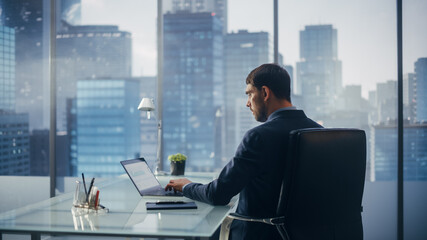 Relaxed Businessman in a Suit Sitting at a Desk in Modern Office, Using Laptop Computer, Next to Window with Big City with Skyscrapers View. Successful Finance Manager Planning Work Projects.