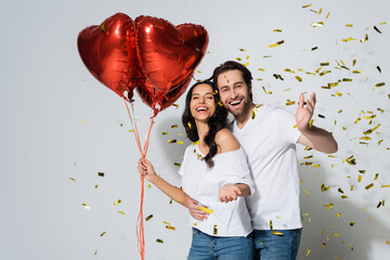 happy woman with red heart-shaped balloons smiling near boyfriend and confetti on grey.