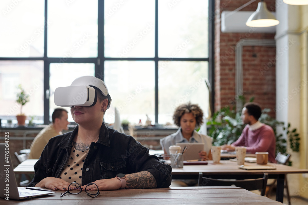 Poster young businesswoman in vr headset sitting by table in front of laptop and preparing presentation in 