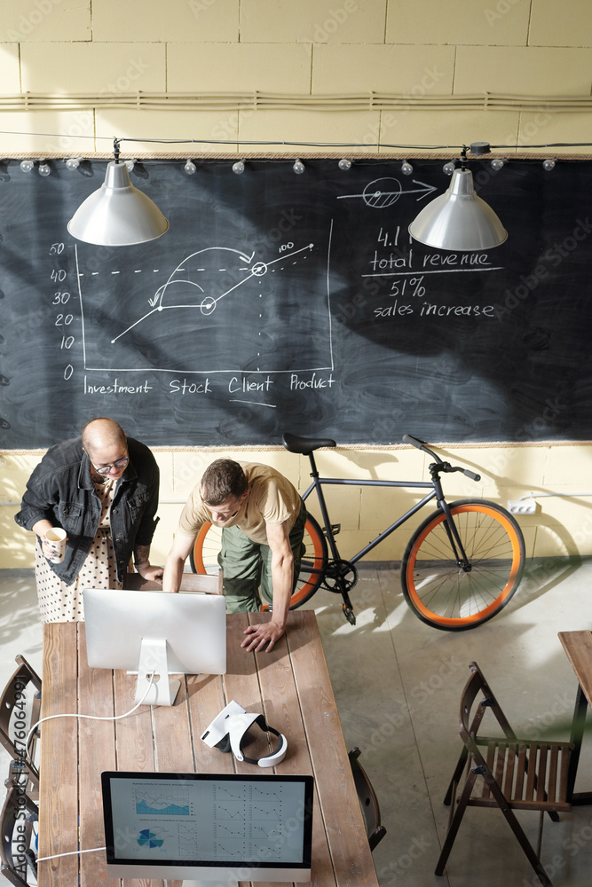 Poster two young caucasian economists bending over table with computer monitor and looking at financial dat