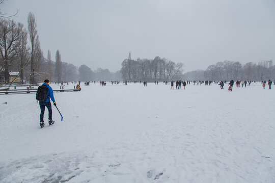 Winter landscape idyllic scene at the frozen lake in the English Garden park, Munich, Germany on a cloudy winter day with people walking, ice skating or playing ice hockey