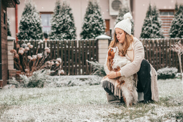 Happy young woman in white knitted hat walks a dog in snowy garden. Cavalier King Charles spaniel. Winter landscape. Pets. Wonderful moments of life. Lot of happiness.Love and care.