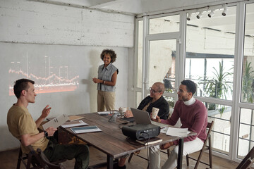 Group of young contemporary economists discussing rates during preentation while gathered by table in office