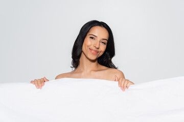 brunette woman smiling at camera while posing behind white blanket isolated on grey.