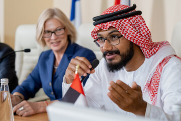 Bearded delegate in turban and eyeglasses making report at international conference while sitting...