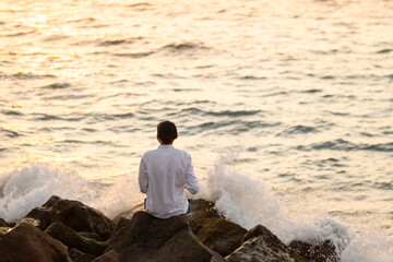 Young fit man in a white linen shirt sitting alone on the rocks by the ocean at sunset hour. Mental health, relaxation, burnout concept.