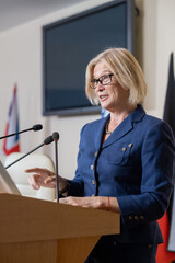 Mature female delegate standing by tribune at political conference and making report for foreign...