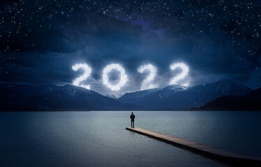 Nieuwjaar 2022 's nachts, man staat op een houten dok aan een meer en kijkt naar de bewolkte nummers in de donkere lucht over bergen, kopieer ruimte