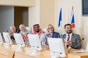 Happy intercultural delegates in formalwear clapping hands to speaker after report while sitting...