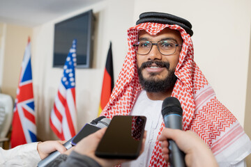 Young confident delegate in national clothes speaking in microphone held by journalist during press...