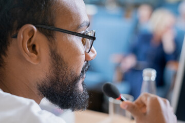 Part of face of young bearded speaker in eyeglasses making speech in front of audience at...