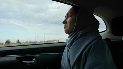 Sad woman wrapped in scarf and in warm coat opens window to breathe fresh air and looks at rainy landscape sitting in comfort car cabin