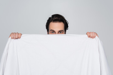 young man looking at camera while obscuring face with white blanket isolated on grey.