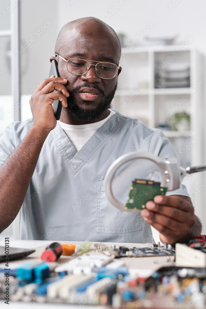 Poster Young confident technician in uniform consulting client on mobile phone while looking at microchip through magnifying glass