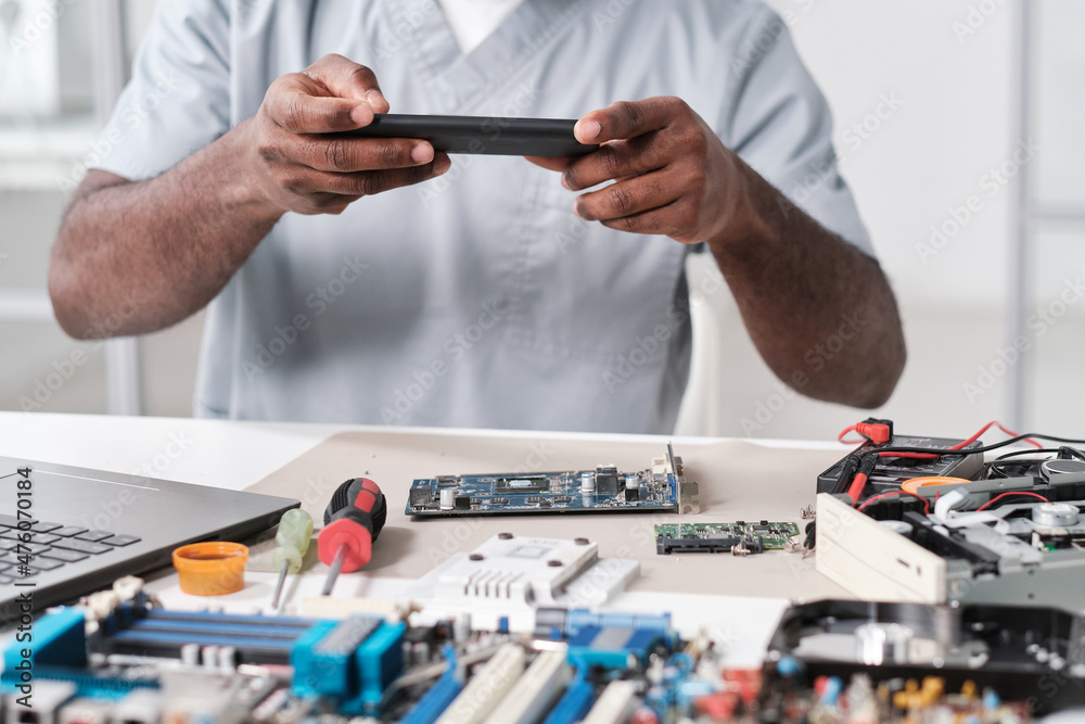 Poster hands of young african repairman photographing circuit board of electronic device while sitting by d