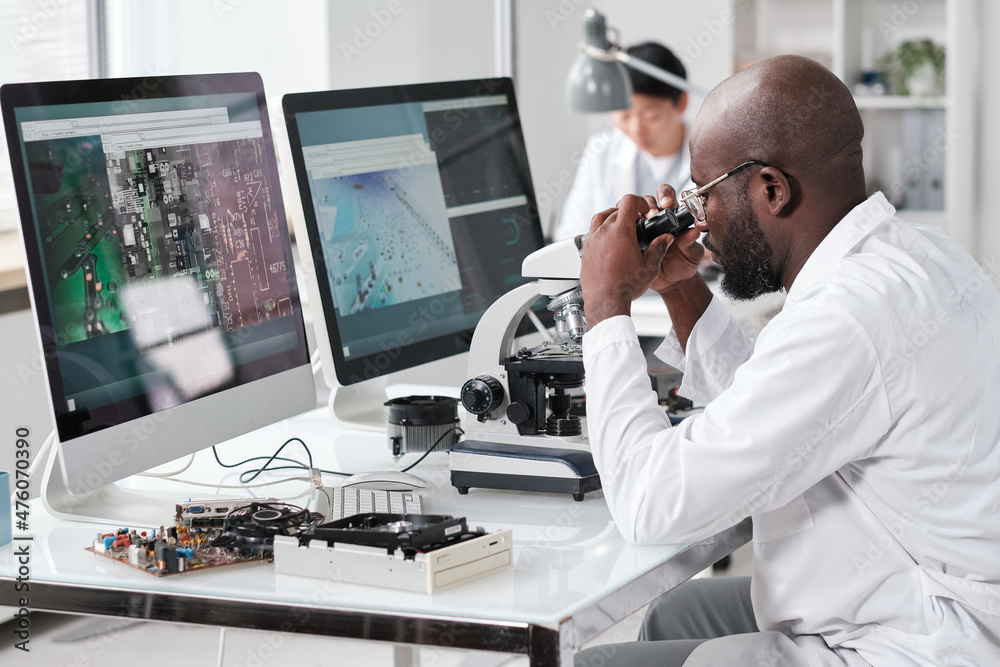 Poster Male laboratory worker in whitecoat studying details of microchip while looking in microscope in front of computer monitors