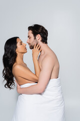 side view of shirtless man and passionate woman in white blanket embracing while standing face to face isolated on grey.