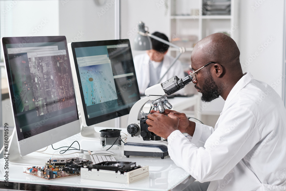 Poster Young serious African man with microscope sitting by workplace in front of computer monitors and colleague on background