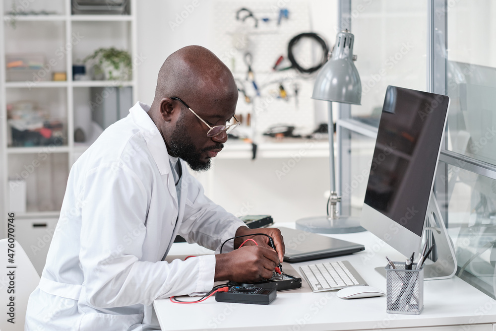 Aufkleber Serious young African repairman in whitecoat soldering microchip in circuit board while bending over desk in front of computer