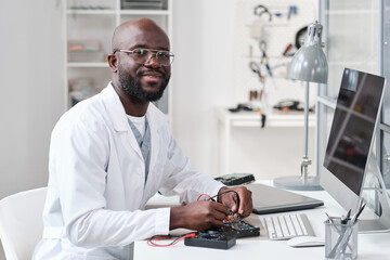 Young cheerful repairman or laboratory worker in whitecoat soldering details of circuit board by workplace and looking at you