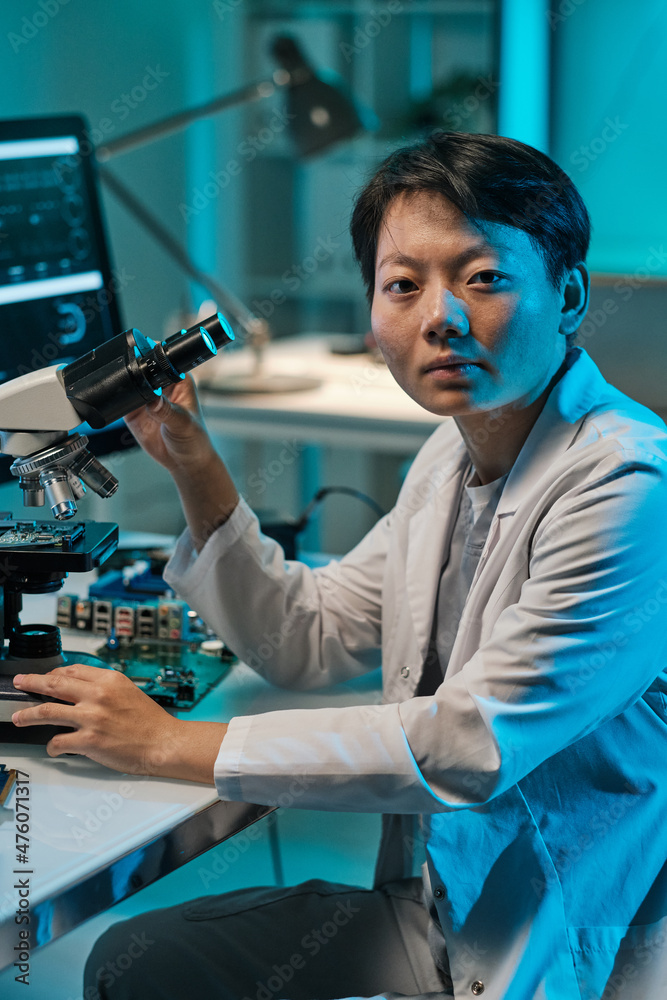 Poster young chinese woman in whitecoat looking at camera while working with microscope in technical labora